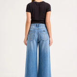 A woman viewed from the back wearing a black short-sleeve t-shirt and high-waisted, wide-leg blue denim jeans against a plain white studio background.