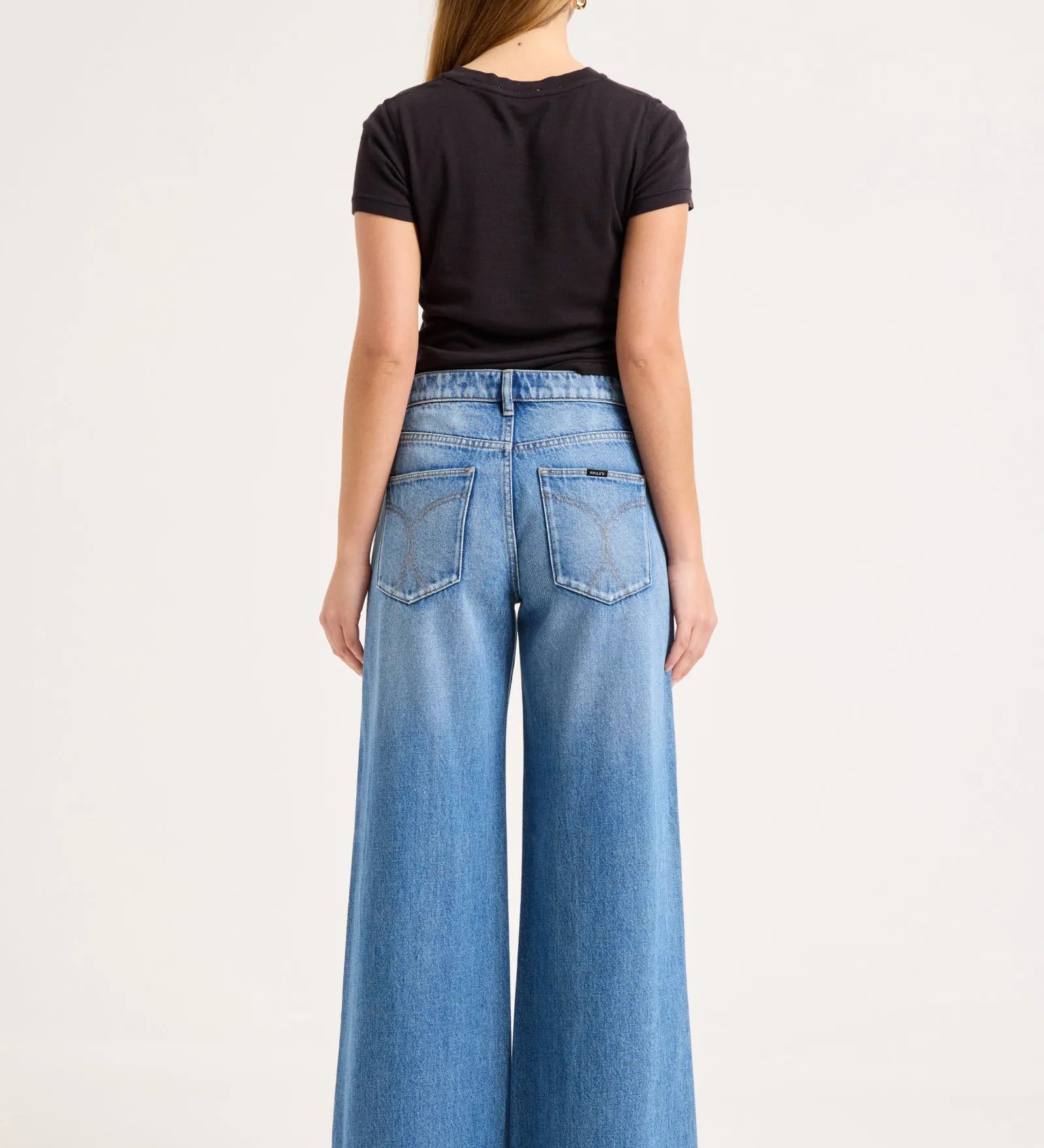 A woman viewed from the back wearing a black short-sleeve t-shirt and high-waisted, wide-leg blue denim jeans against a plain white studio background.