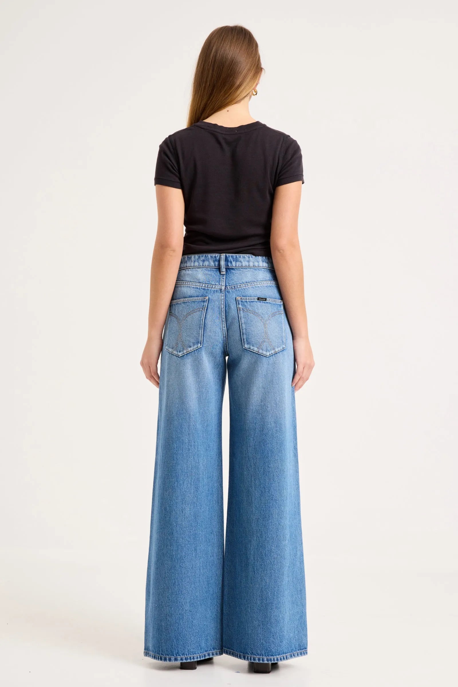 A woman viewed from the back wearing a black short-sleeve t-shirt and high-waisted, wide-leg blue denim jeans against a plain white studio background.