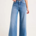 A model wearing a pair of wide-leg blue denim jeans with a black t-shirt and dark pointed shoes, shown from the waist down against a plain light background.
