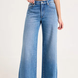 A model wearing a pair of wide-leg blue denim jeans with a black t-shirt and dark pointed shoes, shown from the waist down against a plain light background.