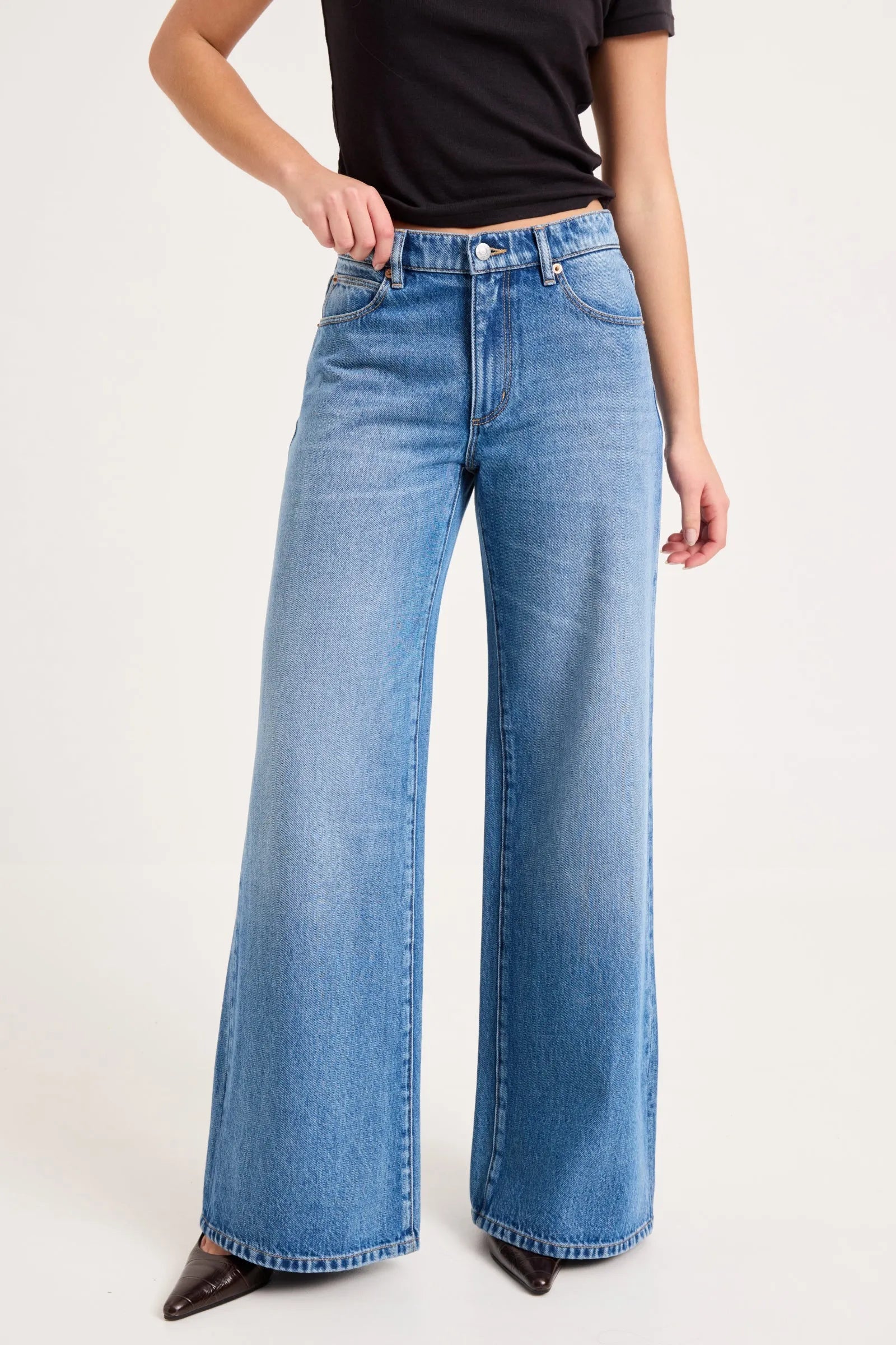 A model wearing a pair of wide-leg blue denim jeans with a black t-shirt and dark pointed shoes, shown from the waist down against a plain light background.