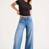 A woman modeling a pair of wide-leg blue jeans with a black short-sleeve crop top, standing against a plain white studio background.