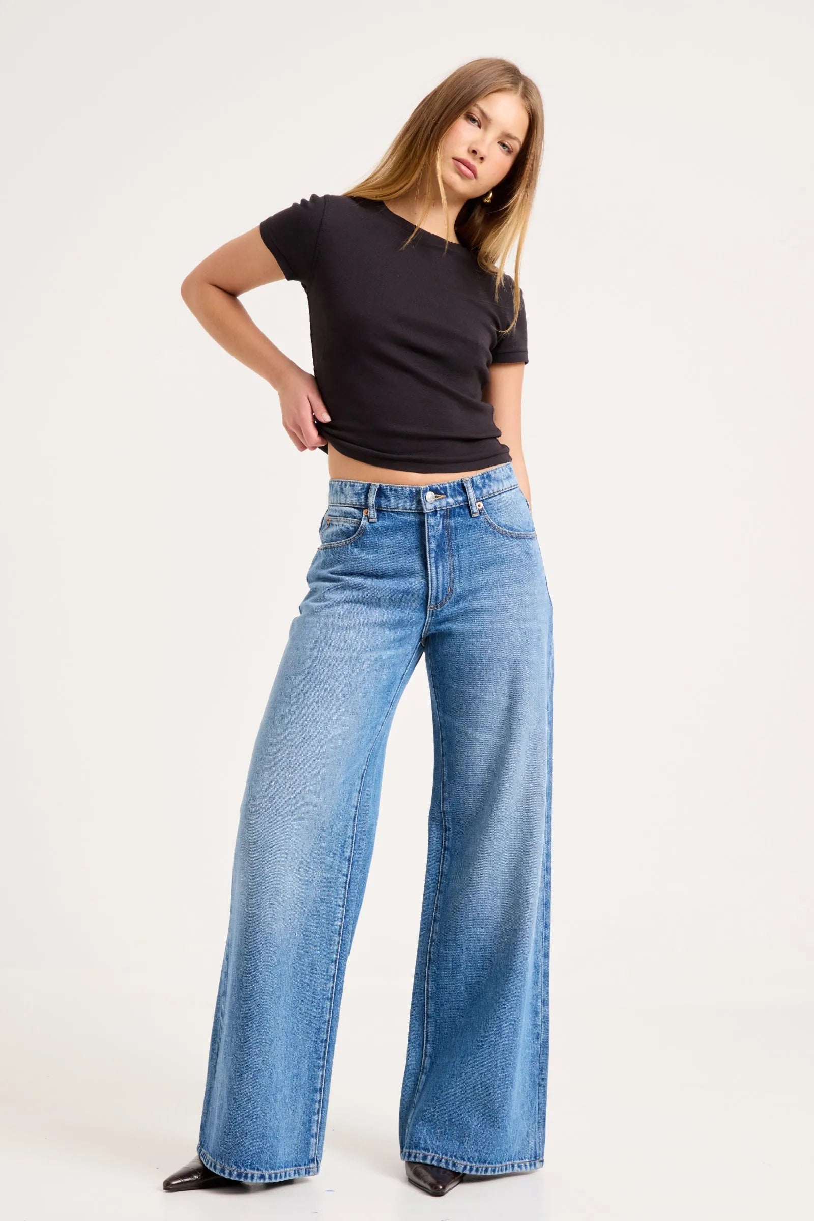 A woman modeling a pair of wide-leg blue jeans with a black short-sleeve crop top, standing against a plain white studio background.