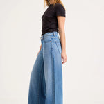 A woman modeling a pair of high-waisted, wide-leg blue denim jeans with a black short-sleeve t-shirt, standing against a plain white studio background.