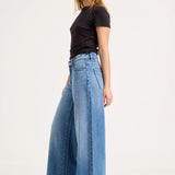 A woman modeling a pair of high-waisted, wide-leg blue denim jeans with a black short-sleeve t-shirt, standing against a plain white studio background.