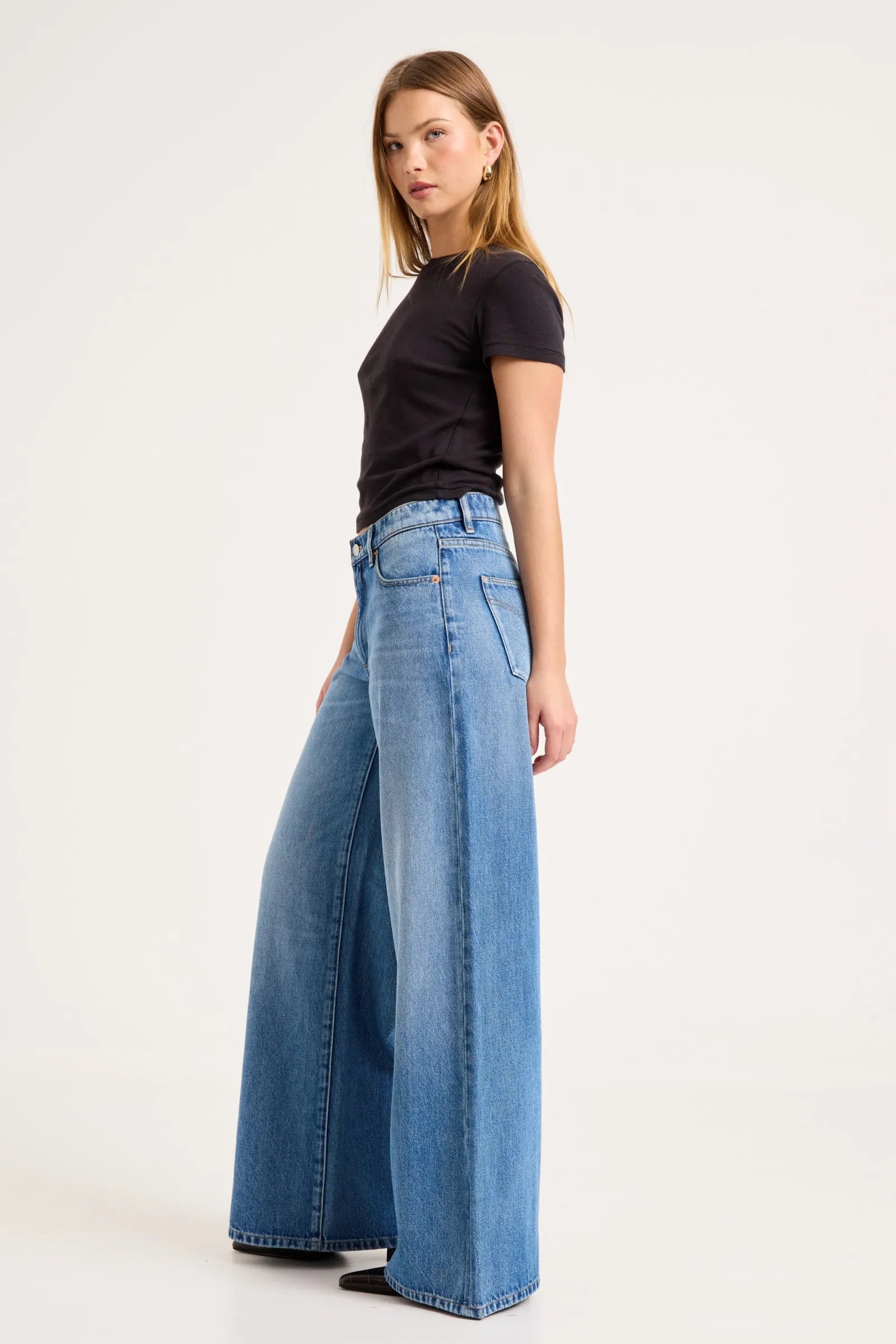 A woman modeling a pair of high-waisted, wide-leg blue denim jeans with a black short-sleeve t-shirt, standing against a plain white studio background.