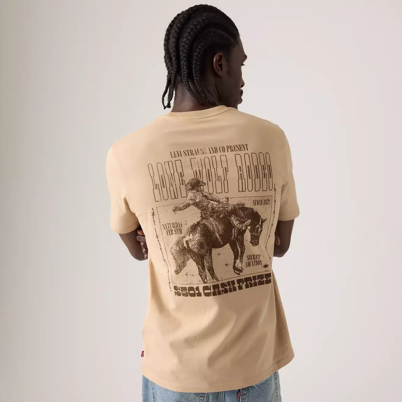 A man shown from behind wearing a beige t-shirt with a large brown graphic print on the back, paired with blue jeans, against a plain light gray background.