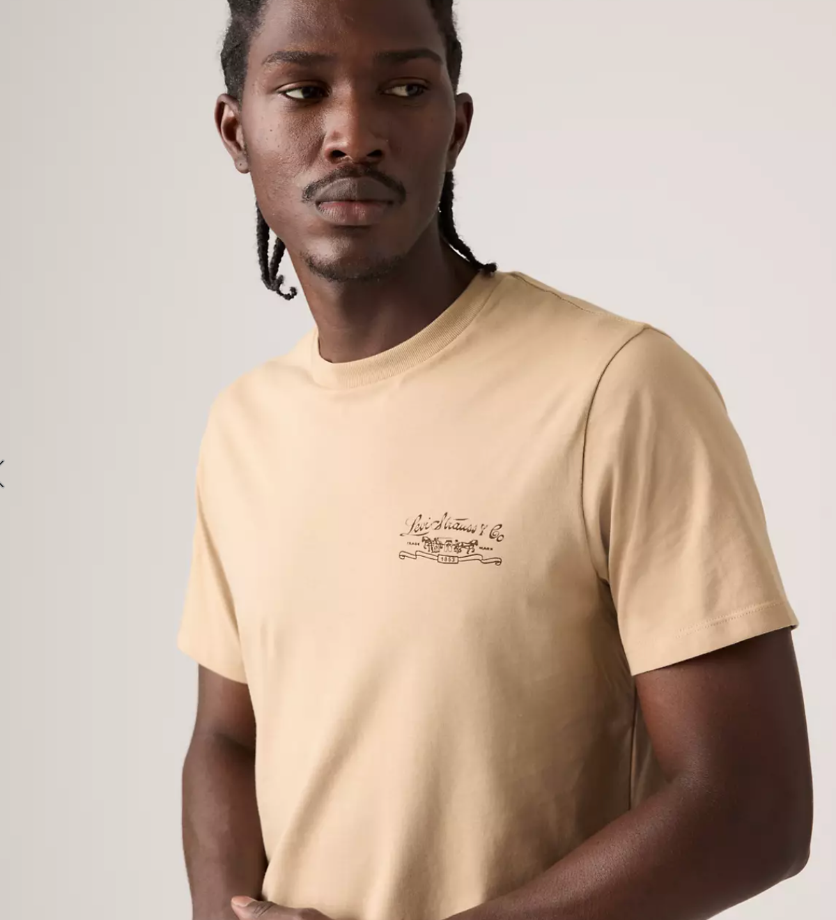 A man wearing a beige crew-neck t-shirt with a small brown script graphic on the left chest, photographed against a light gray studio background.