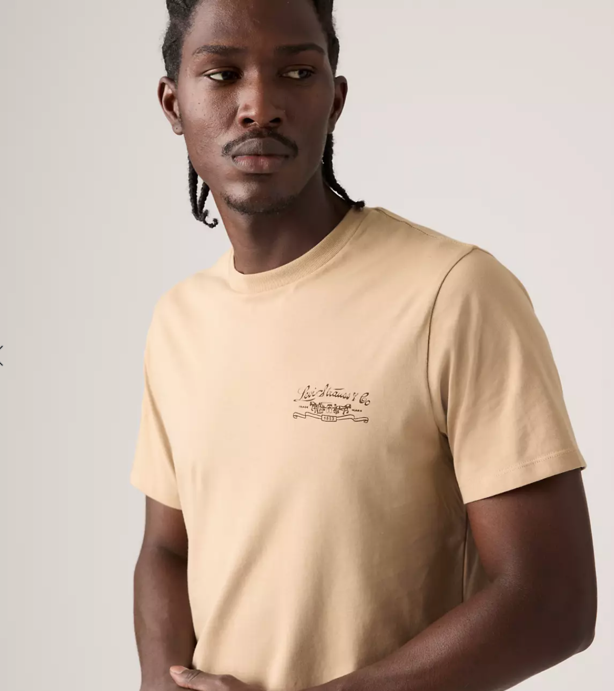 A man wearing a beige crew-neck t-shirt with a small brown script graphic on the left chest, photographed against a light gray studio background.