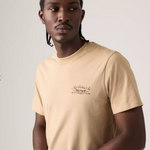 A man wearing a beige crew-neck t-shirt with a small brown script graphic on the left chest, photographed against a light gray studio background.