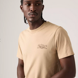 A man wearing a beige crew-neck t-shirt with a small brown script graphic on the left chest, photographed against a light gray studio background.