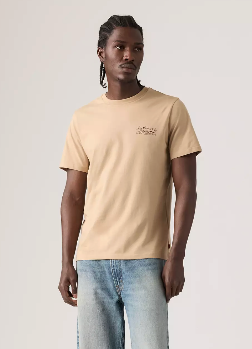 A man wearing a beige short-sleeve crewneck t-shirt with a small graphic on the left chest, paired with light blue jeans, standing against a light gray studio background.