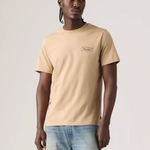 A man wearing a beige short-sleeve crewneck t-shirt with a small graphic on the left chest, paired with light blue jeans, standing against a light gray studio background.