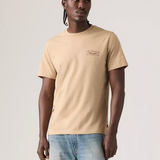 A man wearing a beige short-sleeve crewneck t-shirt with a small graphic on the left chest, paired with light blue jeans, standing against a light gray studio background.