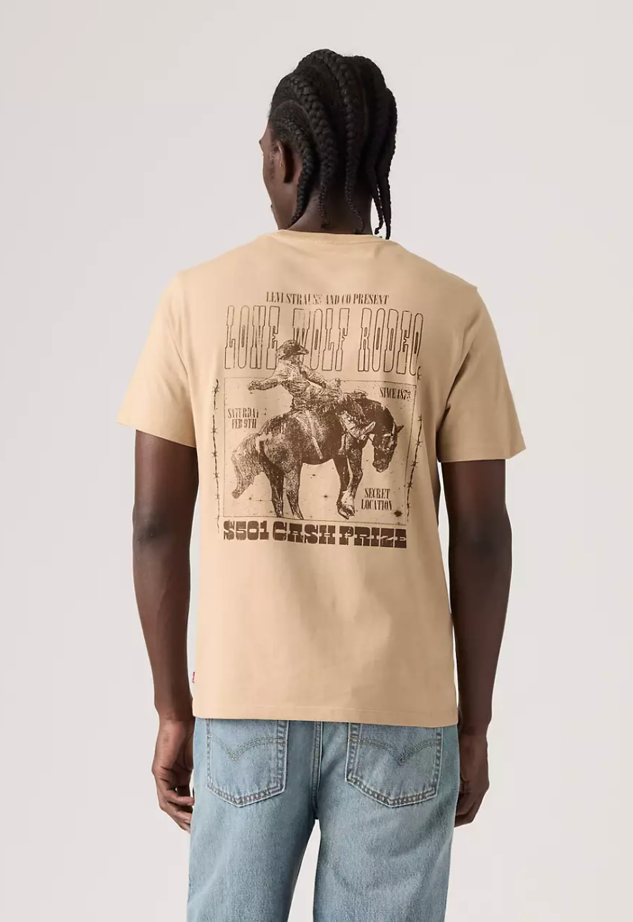 A man viewed from behind wearing a beige graphic t-shirt with a large western-themed print on the back, paired with light blue jeans, against a light gray studio background.