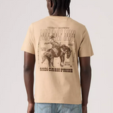 A man viewed from behind wearing a beige graphic t-shirt with a large western-themed print on the back, paired with light blue jeans, against a light gray studio background.