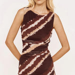 A blonde model wearing a sleeveless ruched mini dress with a brown and beige zebra-like tie-dye pattern, posed against a solid off-white background.