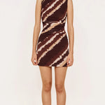 A woman modeling a sleeveless bodycon mini dress with a brown and beige tie-dye pattern, standing against a plain off-white background.