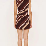 A woman stands with her back to the camera wearing a sleeveless bodycon mini dress with a brown and beige diagonal tie-dye pattern, paired with nude high heels, against a plain off-white studio background.