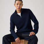Man wearing a navy blue sweater sitting on a wooden block against a plain background