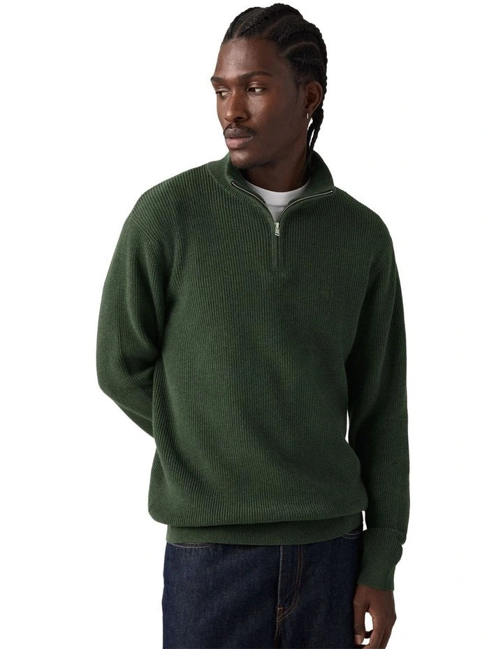 A man wearing a dark green ribbed quarter-zip pullover sweater, paired with dark blue jeans, photographed against a solid white background.