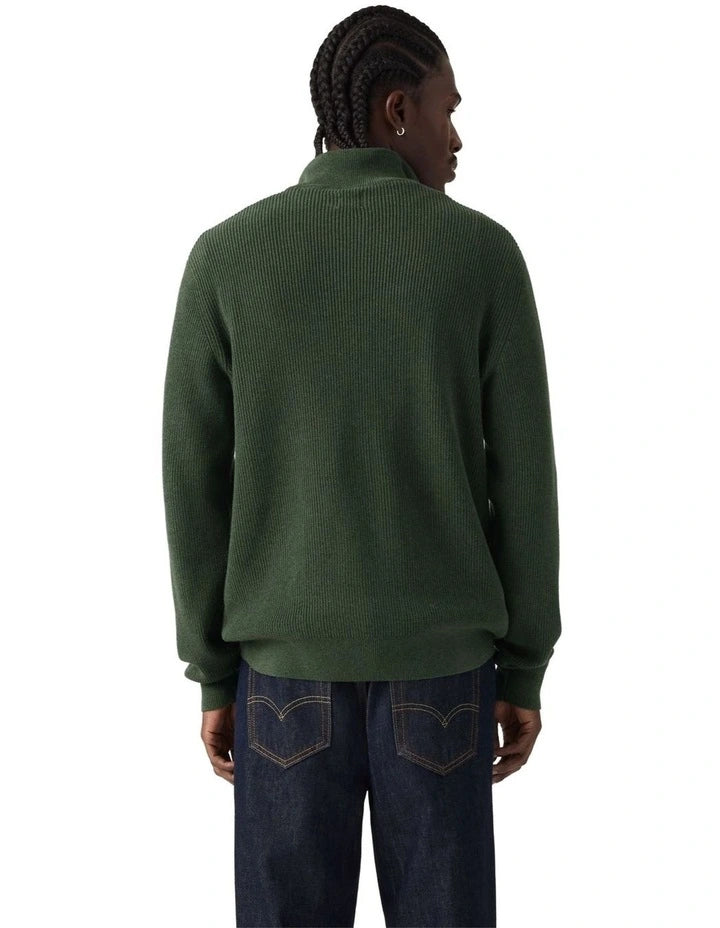 A man viewed from the back wearing a dark green ribbed knit sweater and dark blue jeans against a plain white background.