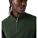 A close-up of a man wearing a dark green ribbed knit quarter-zip pullover sweater against a plain white background.