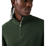 A close-up of a man wearing a dark green ribbed knit quarter-zip pullover sweater against a plain white background.