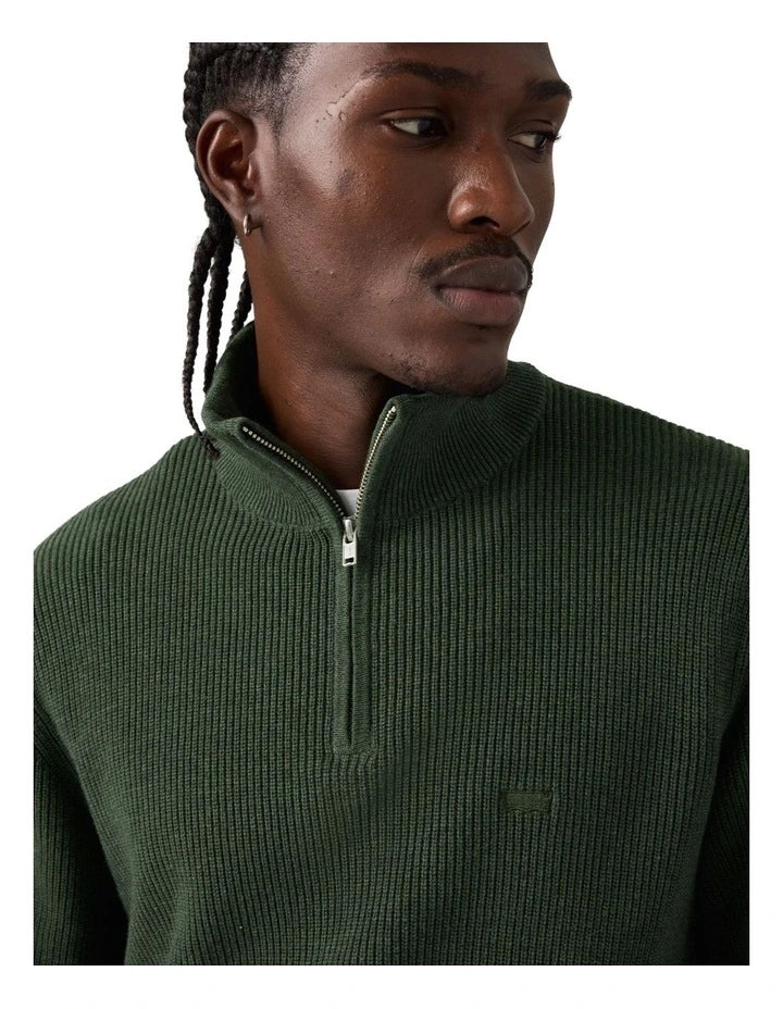 A close-up of a man wearing a dark green ribbed knit quarter-zip pullover sweater against a plain white background.