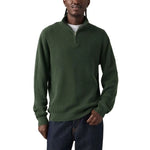 A man wearing a dark green ribbed knit quarter-zip sweater with a small tonal chest logo, paired with dark blue jeans, against a plain white background.