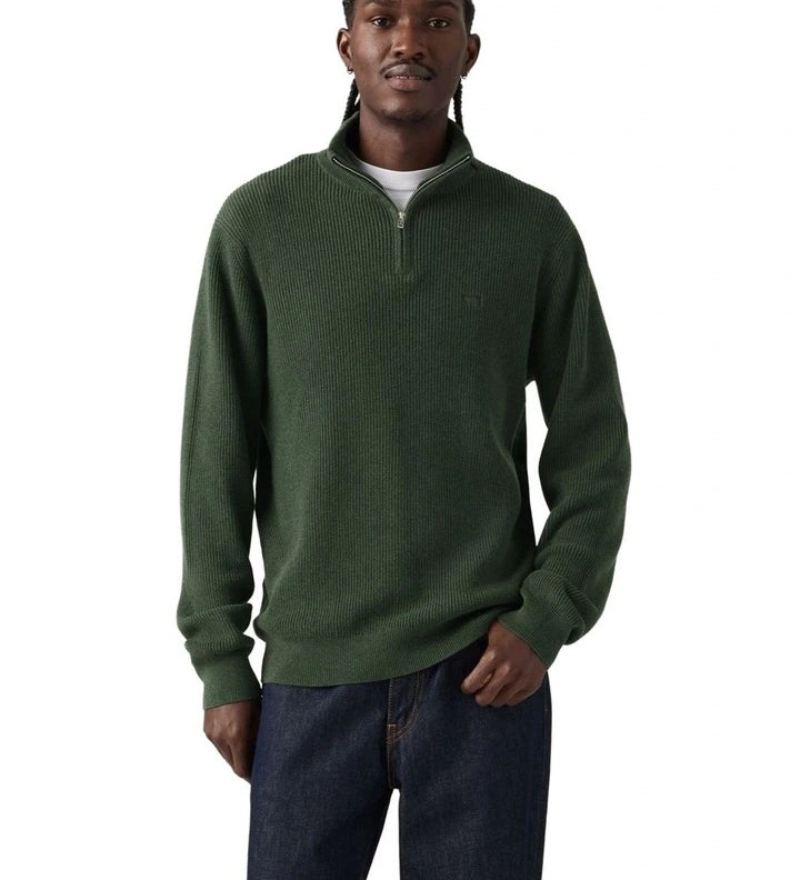 A man wearing a dark green ribbed knit quarter-zip sweater with a small tonal chest logo, paired with dark blue jeans, against a plain white background.