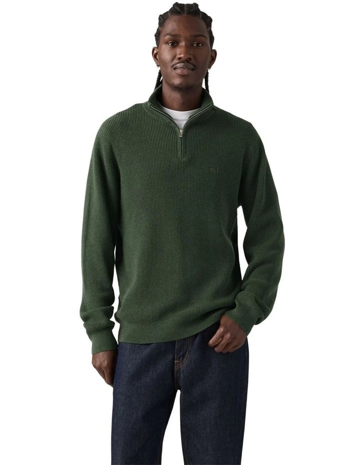 A man wearing a dark green ribbed knit quarter-zip sweater with a small tonal chest logo, paired with dark blue jeans, against a plain white background.