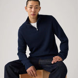 Man wearing a navy blue sweater sitting on a wooden block against a plain background