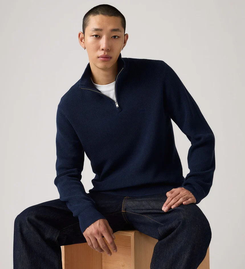 Man wearing a navy blue sweater sitting on a wooden block against a plain background