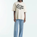 Person wearing a beige t-shirt with a logo and light blue jeans on a white background