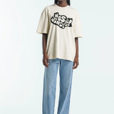 Person wearing a beige t-shirt with a logo and light blue jeans on a white background