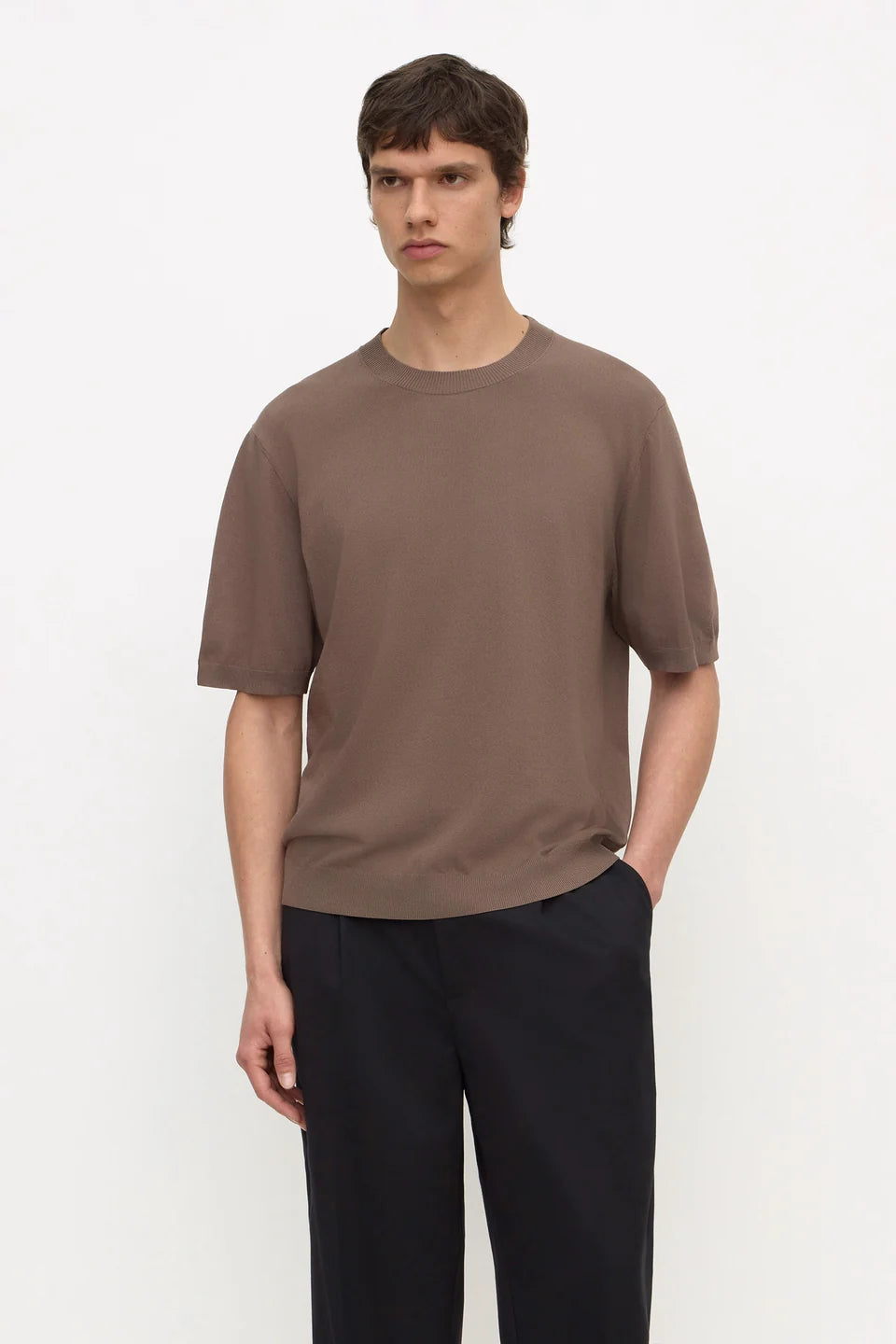 A man wearing a short-sleeve crewneck t-shirt in a muted brown color, paired with dark trousers, standing against a plain white studio background.