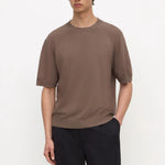 A man wearing a short-sleeve crewneck t-shirt in a muted brown color, paired with dark trousers, standing against a plain white studio background.