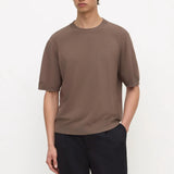 A man wearing a short-sleeve crewneck t-shirt in a muted brown color, paired with dark trousers, standing against a plain white studio background.