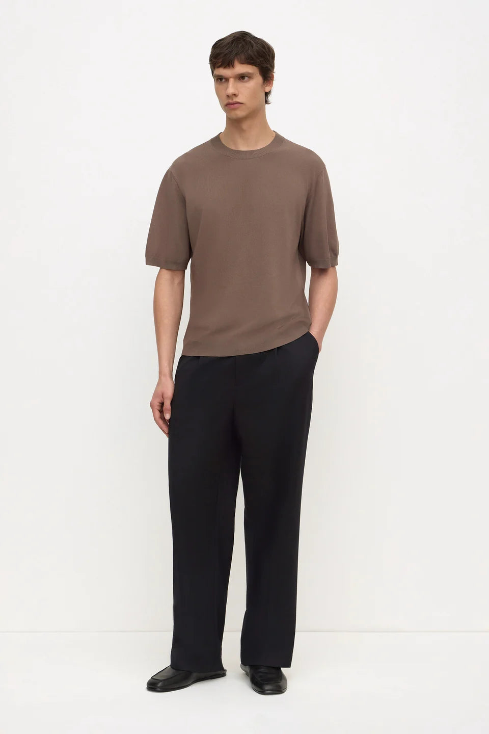 A man modeling a short-sleeve crewneck t-shirt in a muted brown color paired with black wide-leg trousers and black shoes, standing against a plain white studio background.