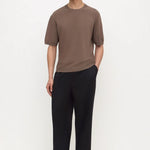 A man modeling a short-sleeve crewneck t-shirt in a muted brown color paired with black wide-leg trousers and black shoes, standing against a plain white studio background.