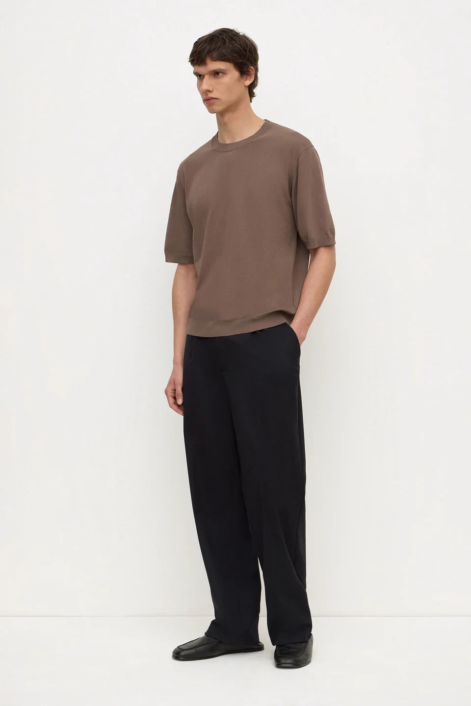 A man modeling a brown short-sleeve crewneck t-shirt and black wide-leg trousers against a plain white studio background.