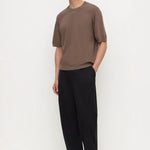 A man modeling a brown short-sleeve crewneck t-shirt and black wide-leg trousers against a plain white studio background.