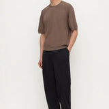 A man modeling a brown short-sleeve crewneck t-shirt and black wide-leg trousers against a plain white studio background.
