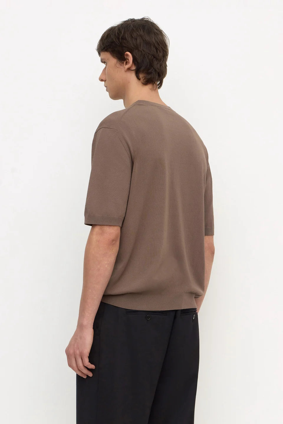 A man wearing a brown short-sleeve knit t-shirt and black trousers, shown from a side-back angle against a plain white studio background.