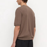 A man wearing a brown short-sleeve knit t-shirt and black trousers, shown from a side-back angle against a plain white studio background.