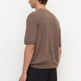 A man wearing a brown short-sleeve knit t-shirt and black trousers, shown from a side-back angle against a plain white studio background.