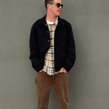 A man wearing a dark denim jacket over a beige and white plaid flannel shirt, paired with brown pants, standing against a textured gray wall.