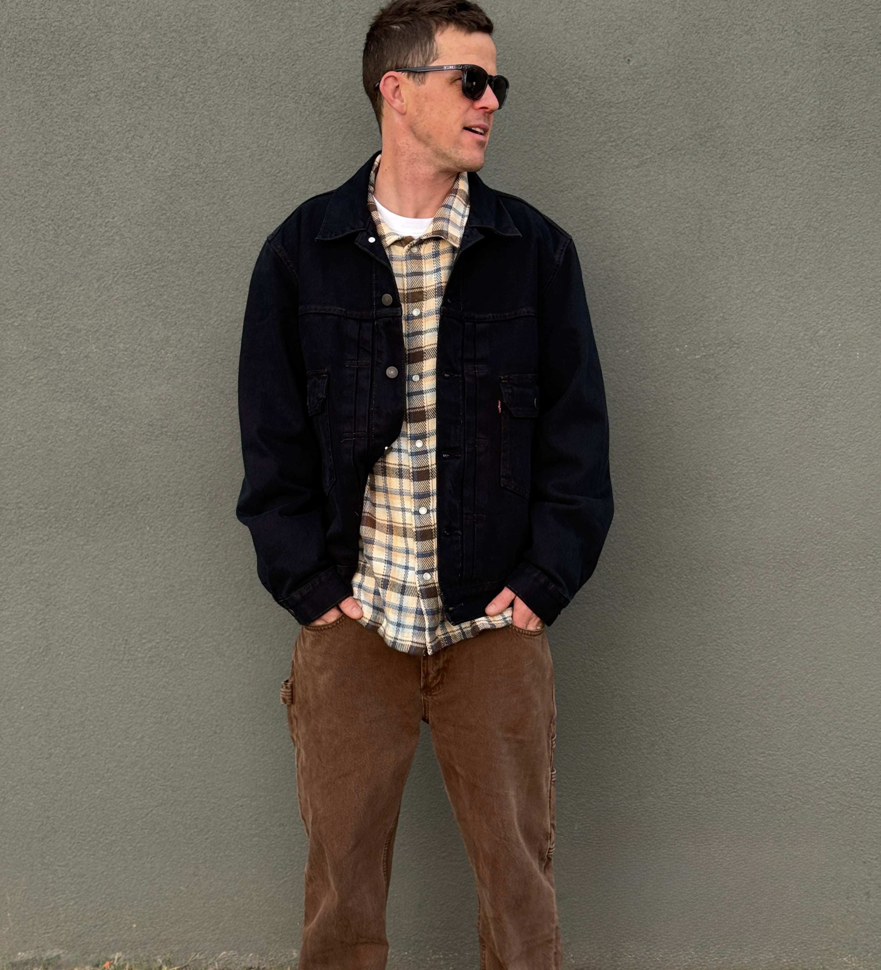 A man wearing a dark denim jacket over a beige and white plaid flannel shirt, paired with brown pants, standing against a textured gray wall.